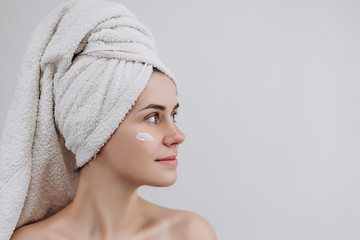 Woman with a towel on her head looking above with face moisturizer near eyes. Causing moisturizer cream onto her face. Skin care and beauty concept. Place for text