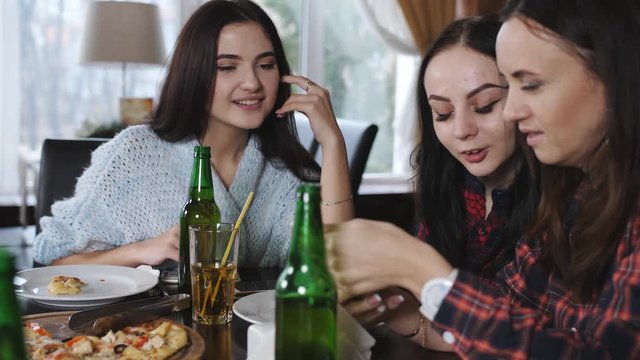 Four girls are relaxing in the restaurant. Drink beer and eat pizza. The girls in the restaurant watch the video on the smartphone and having fun.