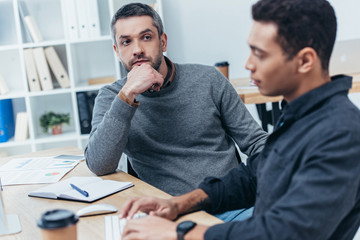 bearded serious business mentor looking at young colleague using desktop computer at workplace