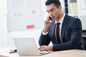 concentrated young businessman talking by smartphone and using laptop at workplace