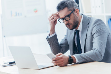 Naklejka premium concentrated businessman in eyeglasses using smartphone and working with laptop in office