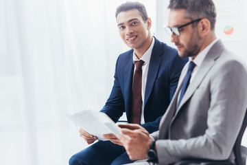 young businessman holding papers and smiling at camera while male colleague in eyeglasses using...