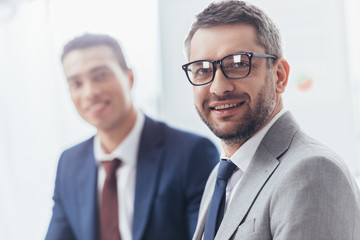 handsome bearded businessman in eyeglasses smiling at camera, young colleague standing behind