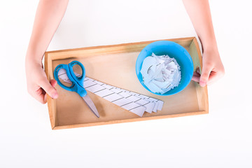 Children's hands hold a wooden tray with montessori materials for a lesson from the practical life zone. Blue scissors, bowl, sheets for cutting. Isolate on white background.
