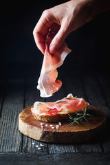 Female hand putting prosciutto on bread over dark wooden background. Sandwich cooking. - obrazy, fototapety, plakaty