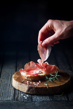 Female hand putting prosciutto on bread over dark wooden background. Sandwich cooking.