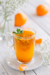 Hot tea with sea buckthorn, fresh mint, mandarin or tangerine lobules on a white wooden table. White porcelain teapot and glass cup. White flowers. 