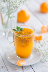 Hot tea with sea buckthorn, fresh mint, mandarin or tangerine lobules on a white wooden table. White porcelain teapot and glass cup. White flowers. 