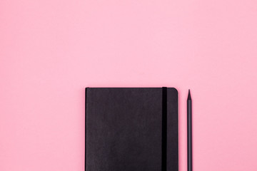 Black closed notebook with pencil on pink background. Minimal and modern freelancer workplace concept. Flat lay, mock up