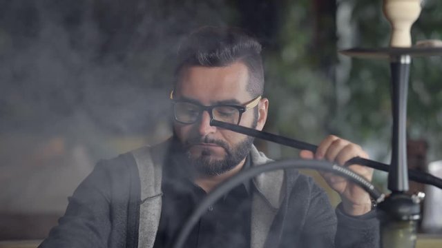 Close-up shot of a stylish adult guy smoking hookah indoor of a cafe alone.