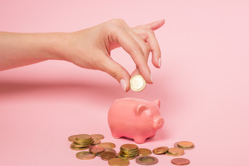Female hand inserting a one euro coin into a pink ceramic piggy bank. Photographed over a pink...