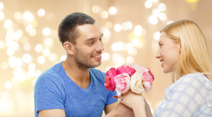 love, couple, greeting and people concept - smiling man giving woman flowers over beige background with festive lights
