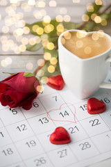 valentines day and holidays concept - close up of calendar sheet with 14th february date, heart shaped chocolate candies, coffee cup and red rose