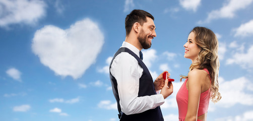 love, couple, proposal and valentines day concept - happy man giving diamond engagement ring in little red box to woman over blue sky and heart shaped cloud background