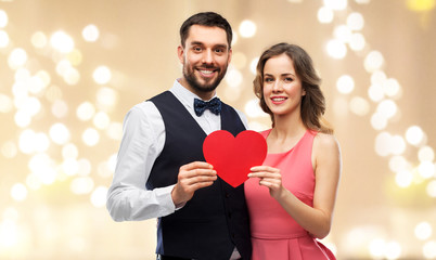Fototapeta na wymiar valentines day, love and people concept - happy couple in party clothes with red heart over festive lights background