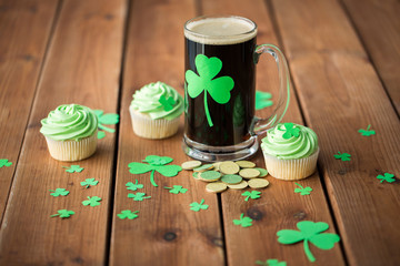 st patricks day, holidays and celebration concept - glass of dark draft beer with shamrock, green cupcakes and gold coins on wooden table