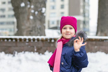 Portrait of happy adorable child showing OK sigh. Cute little caucasian girl in knitted hat and scarf and dawn jacket having fun playing outdoor in winter. Weather and seasons