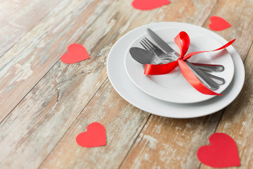 valentines day, table setting and festive dinner concept - plate with spoon, knife and fork tied with red ribbon on wooden background decorated by paper hearts