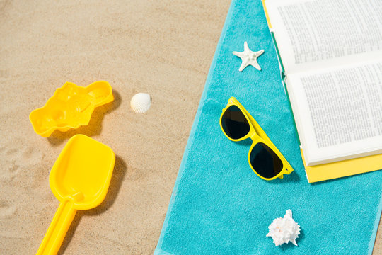 Vacation, Travel And Summer Holidays Concept - Yellow Sunglasses, Sand Toys, Seashells And Book On Beach Towel