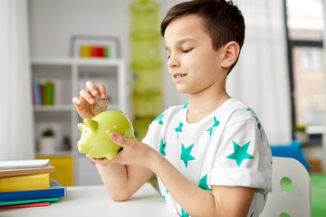 money, finances, childhood and people concept - smiling little boy putting coin into piggy bank at...