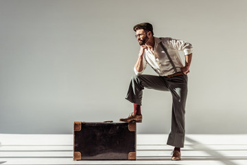 bearded man in glasses and cap with vintage suitcase looking at camera on grey background
