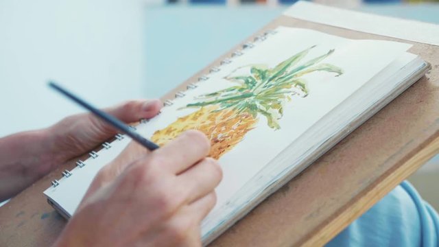 Close-up of female hand with paintbrush is drawing a pineapple by watercolors