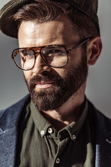 Close up view of handsome bearded man in glasses and cap looking at camera isolated on grey