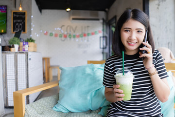 Attractive Young Woman Talking on Mobile Phone at coffee shop  drinking green tea while sitting on the sofa with Happy Facial Expression.