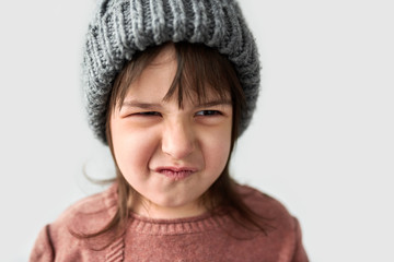 Studio closeup portrait of cute unhappy little girl with grumpy emotion in the winter warm gray hat, wearing sweater isolated on a white studio background.