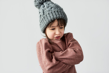 Studio image of pretty angry little girl with grumpy emotion in the winter warm gray hat, wearing sweater isolated on a white studio background.
