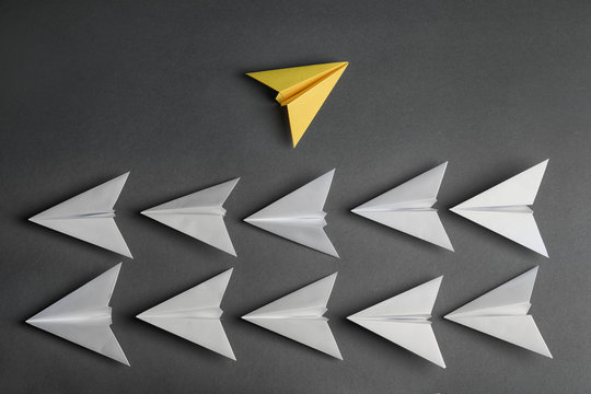 Different color paper plane flying away from others on dark background, top view