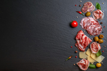 flat lay of antipasti with salami, cheeses, prosciutto, ham, olives, greens, tomato on black slate background, copy space