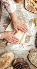 Hands kneading raw dough on table. Top view on baker workplace, working with pastry, all surface on flowered table is occupied with loaves of bread and ingredients. Culinary, cooking, bakery concept