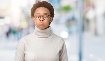 Obraz premium Young beautiful african american woman wearing glasses over isolated background puffing cheeks with funny face. Mouth inflated with air, crazy expression.