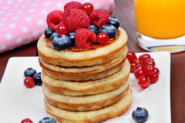 Stacked of pancakes in a plate with fresh berries and orange juice on table
