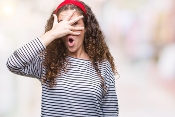 Beautiful brunette curly hair young girl wearing stripes sweater over isolated background peeking in shock covering face and eyes with hand, looking through fingers with embarrassed expression.