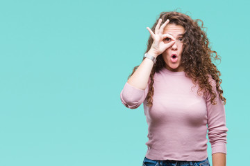 Beautiful brunette curly hair young girl wearing pink sweater over isolated background doing ok gesture shocked with surprised face, eye looking through fingers. Unbelieving expression.
