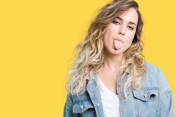 Beautiful young blonde woman wearing denim jacket over isolated background sticking tongue out...