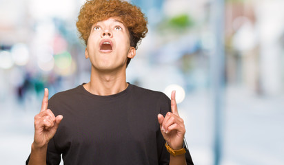 Young handsome man with afro hair wearing black t-shirt amazed and surprised looking up and pointing with fingers and raised arms.