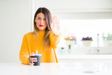 Obraz na płótnie Canvas Young beautiful woman drinking a cup of coffee at home with open hand doing stop sign with serious and confident expression, defense gesture