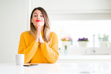 Young beautiful woman drinking a cup of coffee at home praying with hands together asking for forgiveness smiling confident.