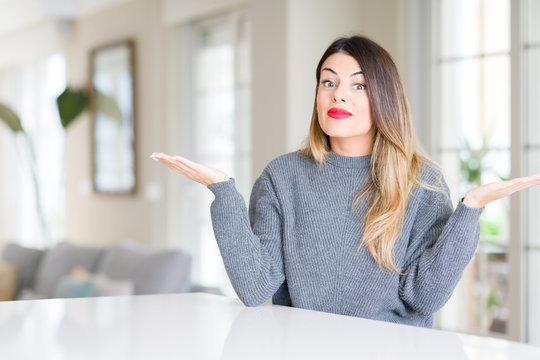 Young beautiful woman wearing winter sweater at home clueless and confused expression with arms and hands raised. Doubt concept.