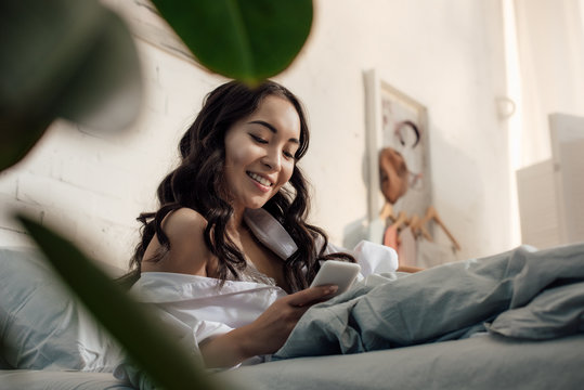 low angle view of beautiful smiling asian girl using smartphone in bed