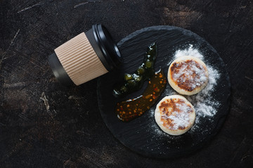 Stone slate with cheesecakes or syrniki and black takeaway coffee cup, flatlay on a dark brown stone background