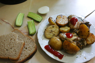 Chicken wings with potatoes lying on the white plate. Homemade lunch. Unhealthy food photo with fat and ketchup tomato sauce. Bread, onion and cucumbers on the beige background.