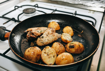 Chicken wings with potatoes lying on the frying pan on the oven. Homemade lunch. Unhealthy food photo with fat.