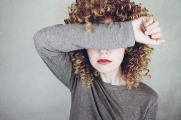 Close-up of a beautiful and young woman with curly blonde hair covers her face with her arm, she...