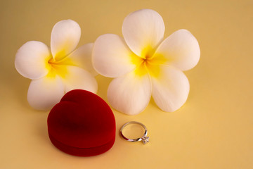 Fototapeta na wymiar Elegant diamond engagement ring and red heart shaped box with tropical white flowers (plumeria) on yellow background. Concept for greeting card, postcard. Copy space.