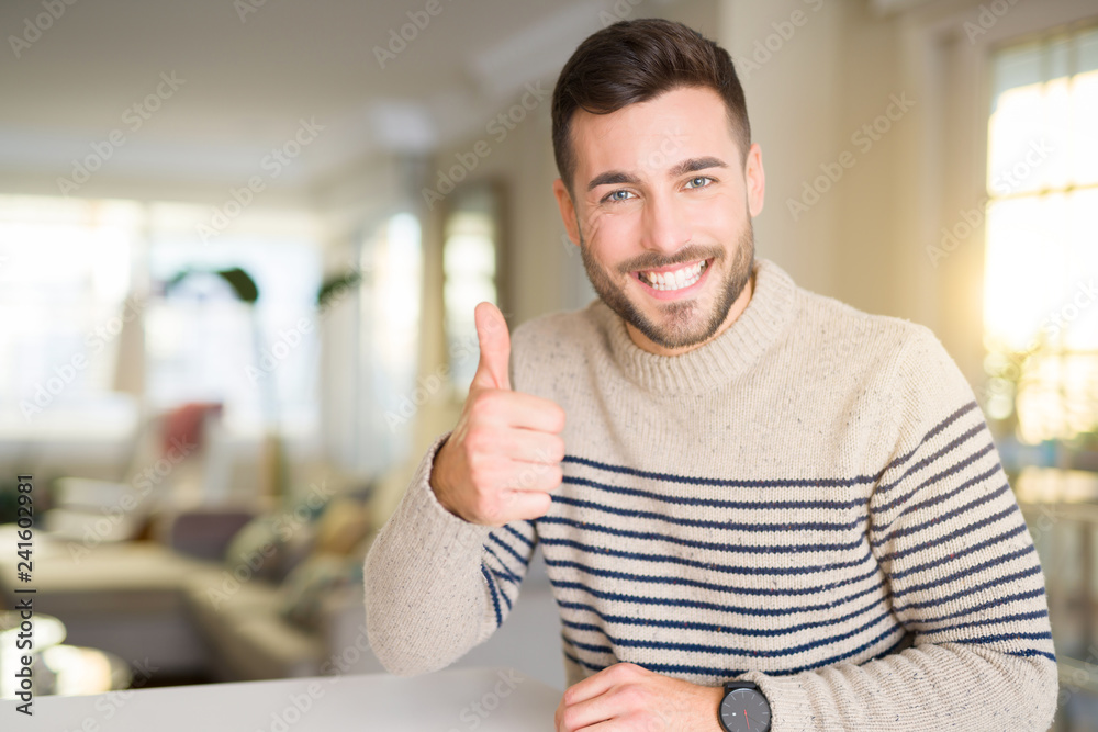 Wall mural young handsome man at home doing happy thumbs up gesture with hand. approving expression looking at 