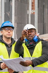 Civil engineers with hardhat and yellow jacket checking technical drawings and office blueprints among scaffolding on construction site